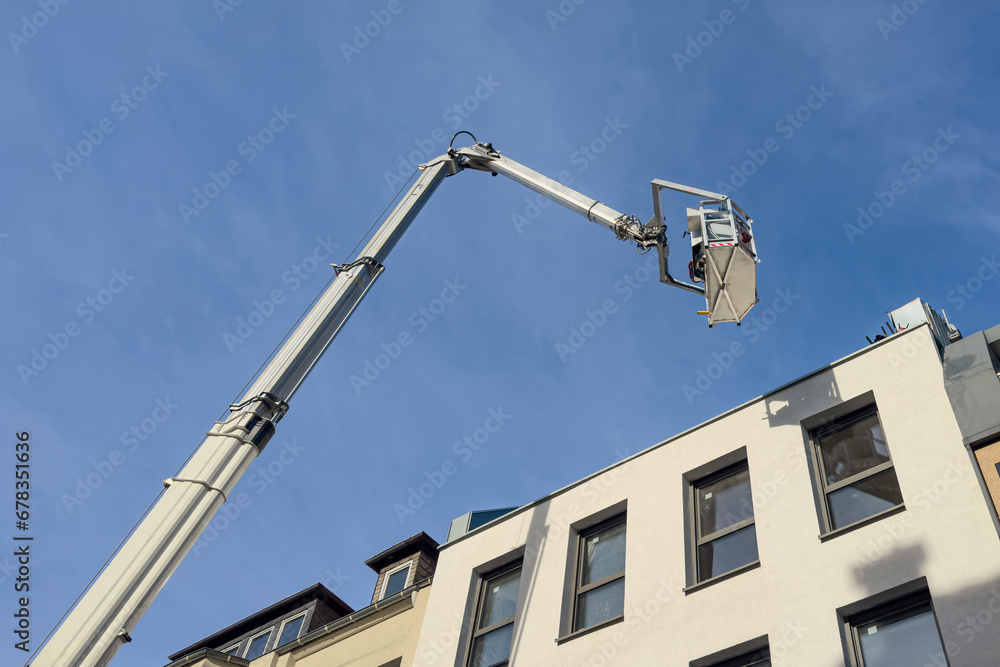 Bottom view of the cabin and boom of a hydraulic lift above a white building against a background of blue sky with light clouds. Bottom view of the cabin and boom of a hydraulic lift above a white building against a background of blue sky with light clouds.