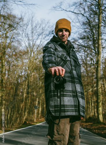 young man on the road holding a vintage camera with the forest behind © Emma