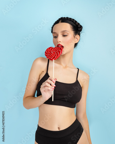 Stylish girl licking lolipop, eating candy and smiling, standing against blue background