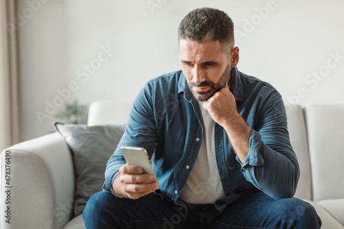 Thoughtful middle aged man doom scrolls through phone at home