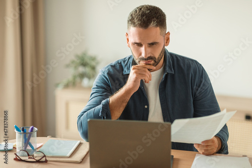 man looking at laptop with thoughtful face holding papers indoor