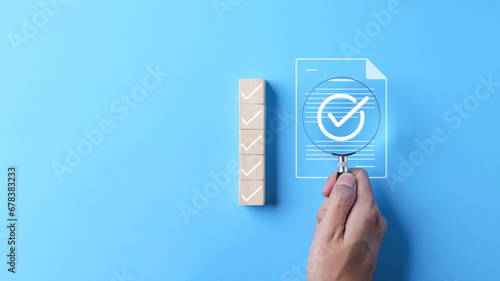Quality assurance document control concept. Businessman holding magnifying glass focus on correct sign mark with document approved paperless and quality assurance approve, Checkmark on wood cube, photo