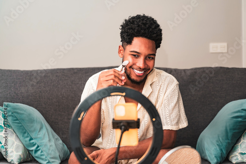 A cheerful young latin man records a skin care theme video blog on his smartphone secured to a ring light, smiling while using a facial roller on a couch at home.