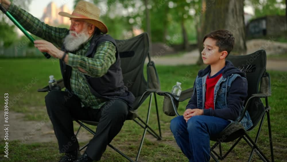 Concept of family relationships. Old angler in a hat with grandson sitting in folding chairs at urban park. Small grandchild gives fish rod to his elderly grandpa. Recreation outdoors together