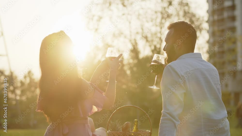 Young family couple having a romantic picnic dinner watching sunset in urban park. Man and woman drinking red wine celebrating first anniversary together outdoors. Date at local lawn Young family couple having a romantic picnic dinner watching sunset in urban park. Man and woman drinking red wine celebrating first anniversary together outdoors. Date at local lawn