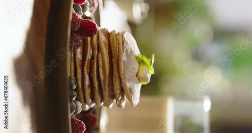 Side view of pancakes adorned with juicy berries, syrup, and sugar powder, kitchen. Leisurely cozy morning, essence of lifestyle, food, cooking, and domestic life. Vertical video, no people, cinematic