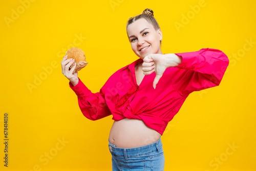 A cute pregnant woman in a pink shirt is eating a fast food burger. A pregnant woman on a yellow background with an appetizing burger in her hands. Harmful food for pregnant women. © Дмитрий Ткачук