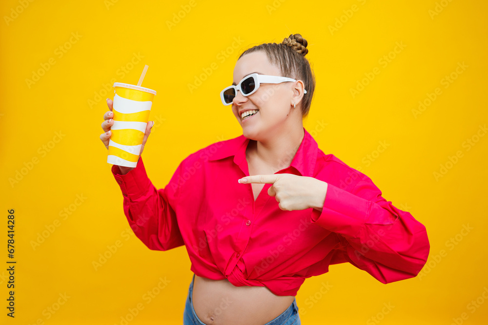 A cute pregnant woman in a pink shirt drinks water from a plastic cup. A pregnant woman on a yellow background with a glass in her hands. Harmful water for pregnant women. A cute pregnant woman in a pink shirt drinks water from a plastic cup. A pregnant woman on a yellow background with a glass in her hands. Harmful water for pregnant women.