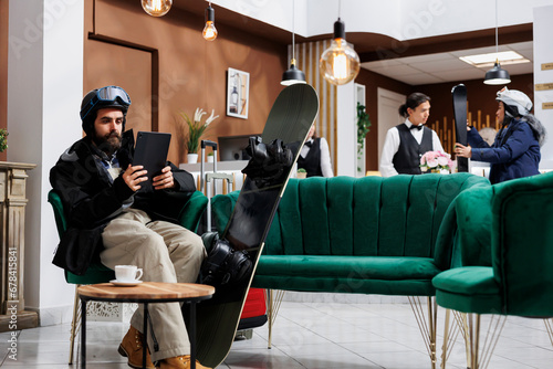 A male guest in winter clothing sits on cozy couch in a hotel reception, using phone tablet to browse and make reservations. Snowboarding gear is nearby, reflecting his enthusiasm for wintersports.