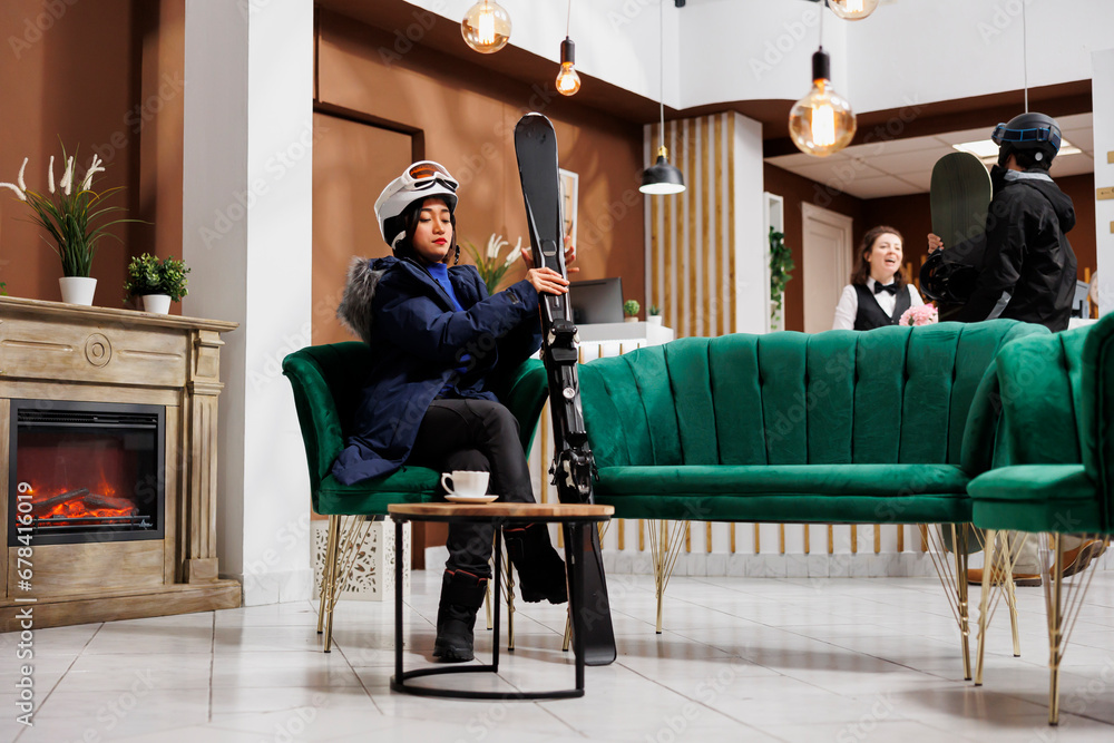 Female traveler seen in lounge area fully equipped for wintersports with snow gear and winter jacket. Woman holding skiing skis seated on sofa while man with snowboard arrives at front desk.