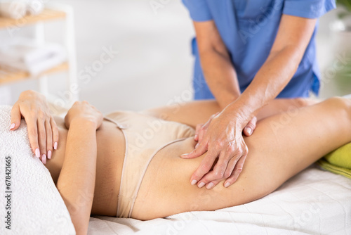Cropped woman dressed in tango panties lies on massage table during spa procedure and receives massage of leg things area. Select focus hand of mature female masseuse