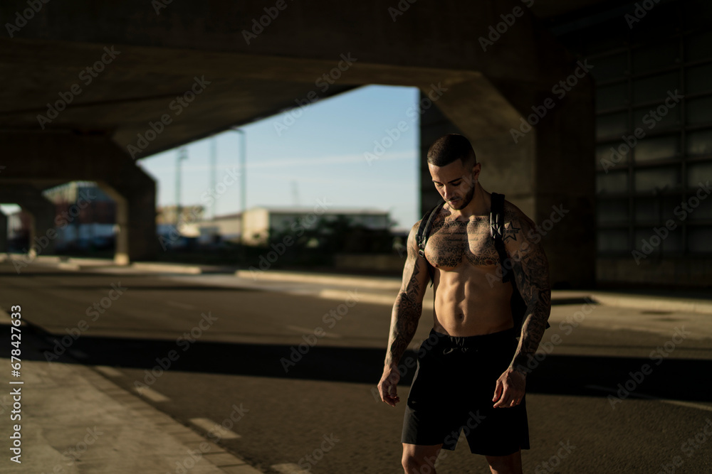 Chico joven tatuado y musculoso posando y haciendo deporte en la calle en un tunel con un puente de fondo