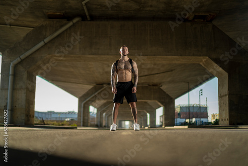 Chico joven tatuado y musculoso posando y haciendo deporte en la calle en un tunel con un puente de fondo