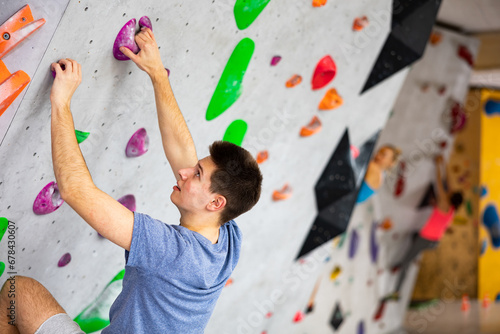 European young man grabbing ledges of artificial climbing wall in bouldering centre.