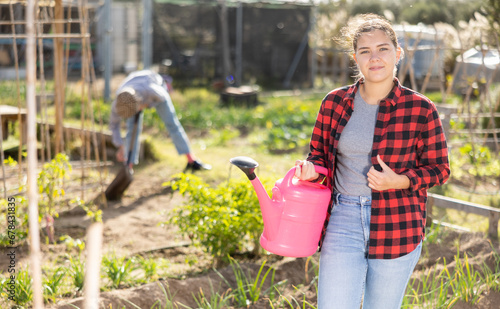 Skilled young woman farmer in plaid shirt holding pink watering can in hands and smiling at camera during work in vegetable garden in autumn © JackF