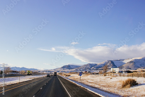 A beautiful landscape with mountains covered in snow, a highway with cars driving along it, and a blue sky with fluffy clouds on a sunny winter day. Butte, Montana, USA