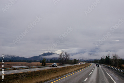 Asphalt road in the middle of high mountains, covered with fog and clouds, at dusk. American winter landscape of a mountainous area covered with fir forest © Liudmila