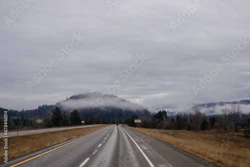 Asphalt road in the middle of high mountains, covered with fog and clouds, at dusk. American winter landscape of a mountainous area covered with fir forest © Liudmila