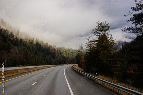 Asphalt road in the middle of high mountains, covered with fog and clouds on autumn day. American winter landscape of a mountainous area covered with fir forest. Fall season on the highway with cars