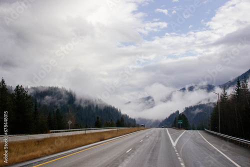 Asphalt road in the middle of high mountains, covered with fog and clouds, at dusk. American winter landscape of a mountainous area covered with fir forest