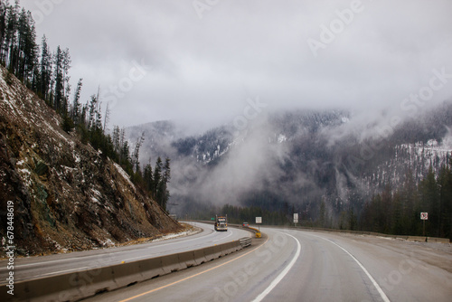 Asphalt road in the middle of high mountains, covered with fog and clouds, at dusk. American winter landscape of a mountainous area covered with fir forest