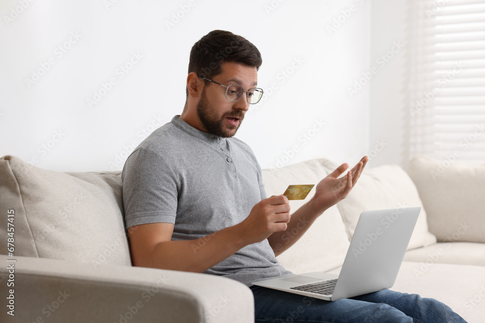 Emotional man with credit card and laptop on sofa at home. Be careful - fraud