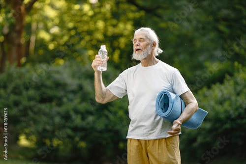 Senior man with exercise mat standing at park and holding bottle of water