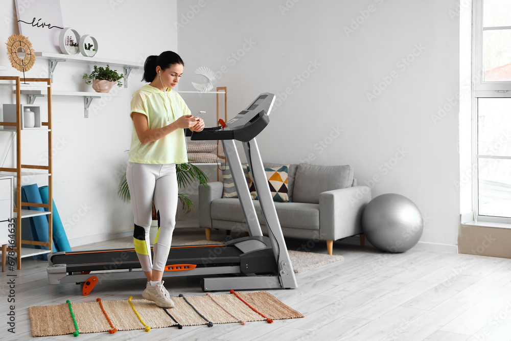 Beautiful woman with earphones using mobile phone near treadmill at home