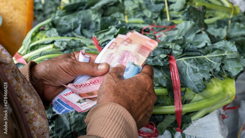 an old woman's hand holding a rupiah note with a denomination of Rp.100,000 against a background of green broccoli vegetables