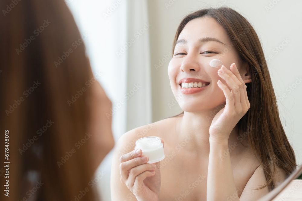 Facial beauty skin care, smile of pretty asian young woman in bathrobe looking at mirror, hand applying moisturizer lotion on her face, holding jar of skin cream before makeup cosmetic routine at home Facial beauty skin care, smile of pretty asian young woman in bathrobe looking at mirror, hand applying moisturizer lotion on her face, holding jar of skin cream before makeup cosmetic routine at home