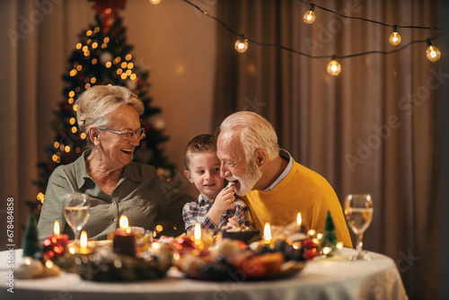 A grandson is feeding grandfather with cookies at christmas table while sitting together with grandmother at home on new year and christmas eve.
