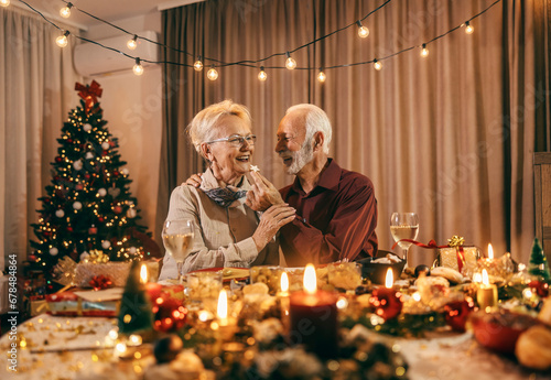 Fototapeta A senior man is feeding his wife with cookie at christmas table on christmas and new year's eve at home