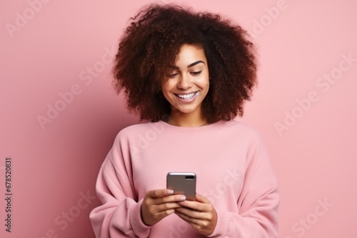 Studio portrait of beautiful African American woman with smartphone in pink clothes against pink background. Positive girl with Afro haircut texting message, enjoying online communication, using app.