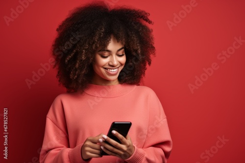 Studio portrait of beautiful African American woman with smartphone in pink clothes against red background. Positive girl with Afro haircut texting message, enjoying online communication, using app.