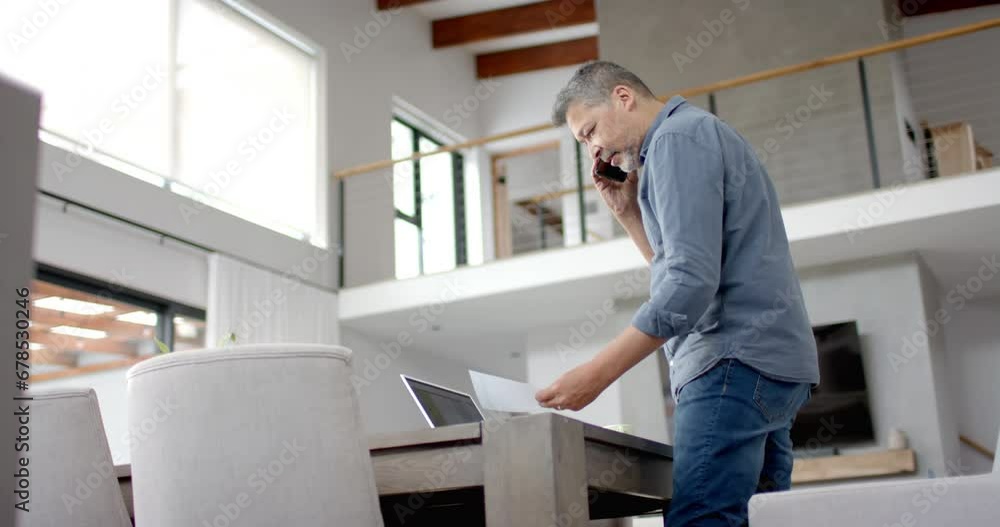 Senior biracial man on phone, standing looking at paperwork, home working, copy space, slow motion Senior biracial man on phone, standing looking at paperwork, home working, copy space, slow motion