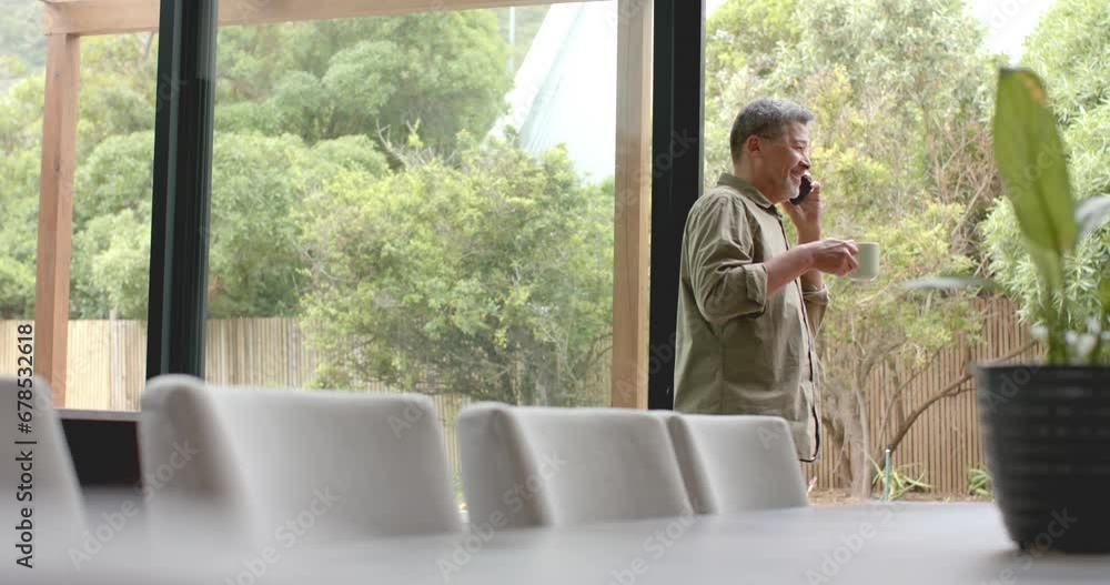 Happy senior biracial man by window talking on smartphone holding coffee, copy space, slow motion Happy senior biracial man by window talking on smartphone holding coffee, copy space, slow motion