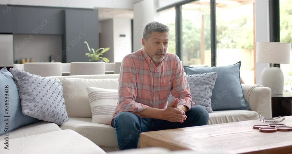Worried senior biracial man sitting on couch thinking in living room, copy space, slow motion Worried senior biracial man sitting on couch thinking in living room, copy space, slow motion