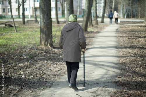 Old lady with walking stick walks through city. Pensioner in park. Old man in town. Walk along path.