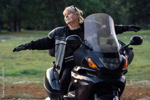 Towards adventures. Happy mature blonde motorcyclist has her arms outstretched, sitting on bike.