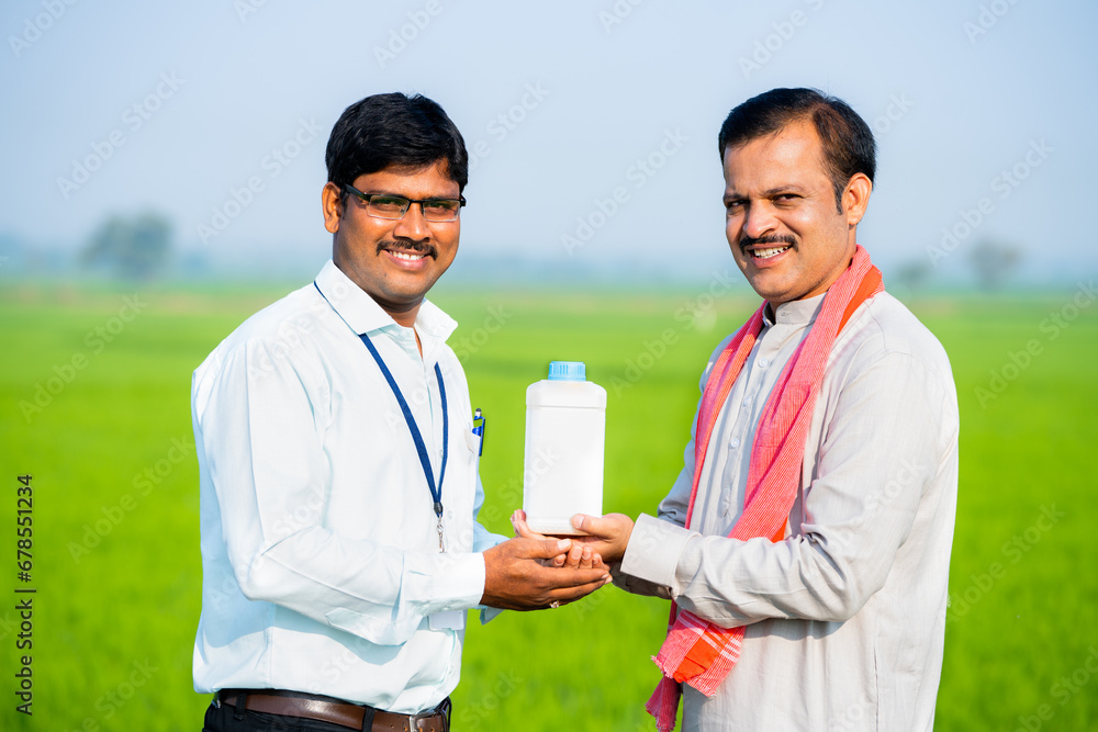 Happy indian agro scientist or officer giving fertilizer to village farmer by looking at camera at farmland - concept of recommendation, satisfied and farming. Happy indian agro scientist or officer giving fertilizer to village farmer by looking at camera at farmland - concept of recommendation, satisfied and farming.