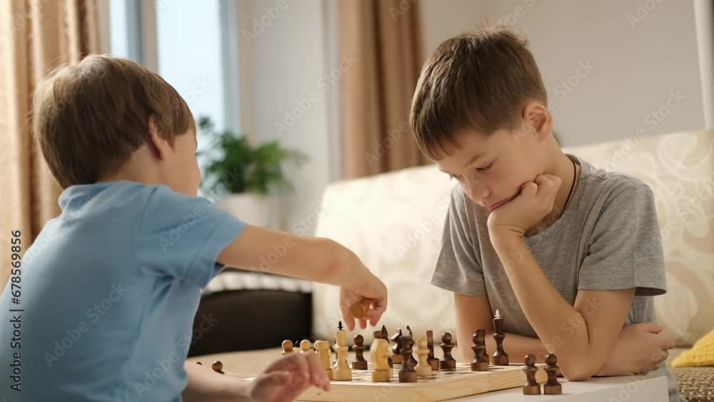 Two Caucasian boys are playing chess at home in living room. Older brother teaches younger one to play chess. Leisure game. Two Caucasian boys are playing chess at home in living room. Older brother teaches younger one to play chess. Leisure game.