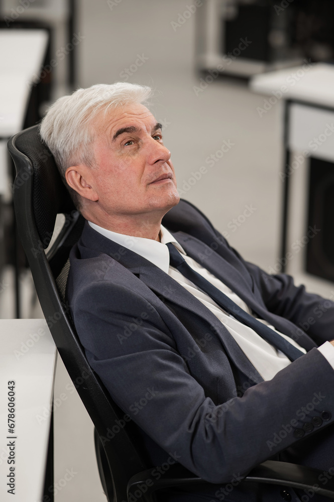Portrait of a pensive mature business man sitting in a chair in the office. Vertical photo.