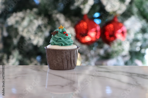Cupcakes decorated with green buttercream. It is a Christmas tree pattern, which has a gold star atop, in a brown cup. Placed on out focus background of Christmas trees with white snow.