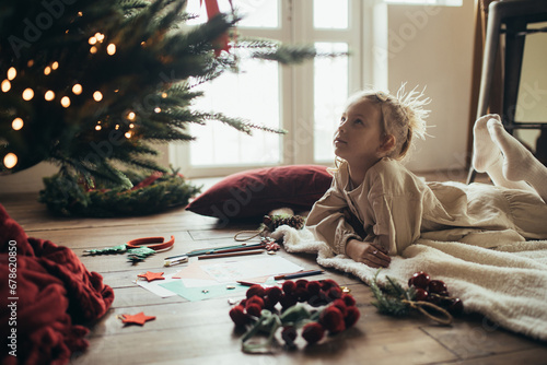 Cute girl writing a letter to Santa Claus, lying on a wooden floor under decorated Christmas tree. photo