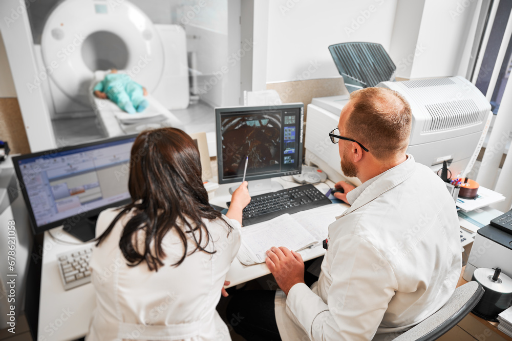 Back view of two doctors sitting, working at computer, examining MRI results. Specialists wearing white robes, consulting, woman pointing by pen, patient lying. Concept of medicine. Back view of two doctors sitting, working at computer, examining MRI results. Specialists wearing white robes, consulting, woman pointing by pen, patient lying. Concept of medicine.