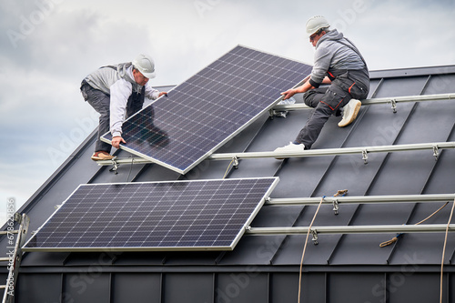 Workers building solar panel system on roof of house. Men technicians in helmets carrying photovoltaic solar module outdoors. Concept of alternative and renewable energy. photo