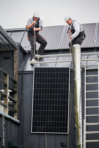 Men electricians installing solar panel system on roof of house. Roofers in helmets lifting up photovoltaic solar module with help of ropes outdoors. Concept of alternative and renewable energy.