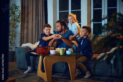 Father spending time with children, sons and daughter, sitting on couch at home and watching tv. Loving and caring parent. Concept of family, leisure time, relaxation, childhood and parenthood