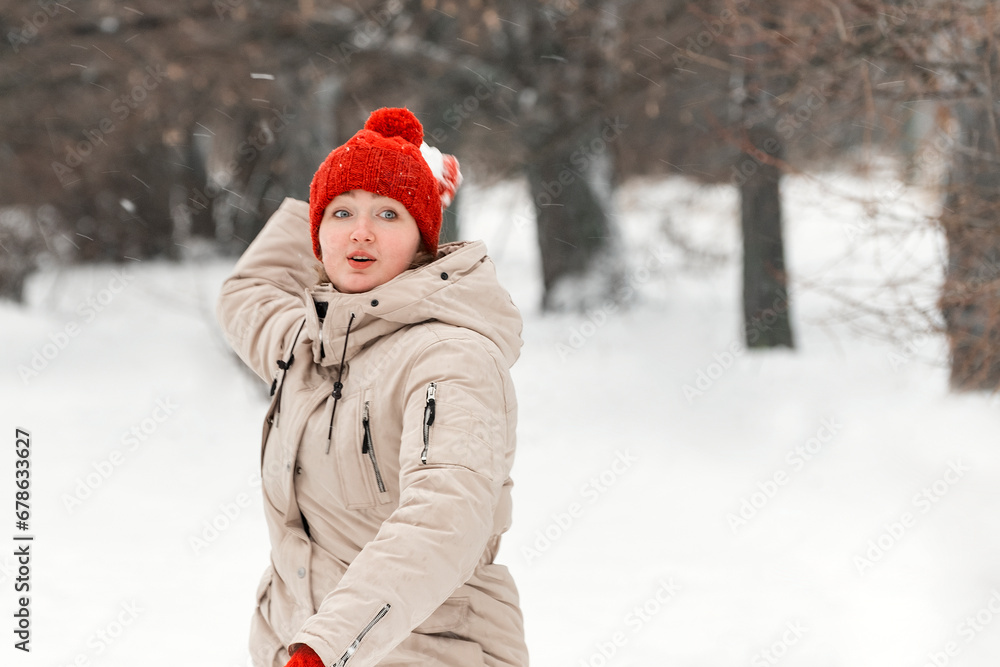 Girl throws snowball walking in winter forest. Young woman in red hat has fun on snow-covered park background
