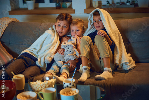 Emotional children, boys and little girl sitting on couch at home and watching tv, horror movie. Hiding under blanket. Concept of family, leisure time, relaxation, childhood and parenthood